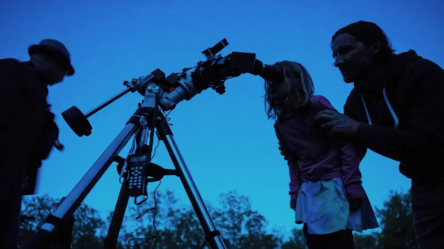 A man helps a child see through the viewport of a telescope against a dark blue dusky sky