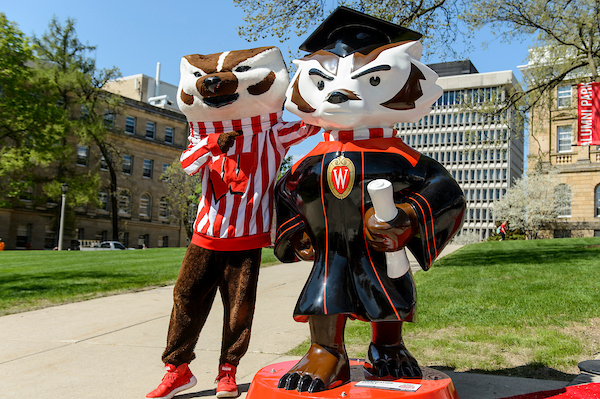UW mascot Bucky Badger stands with his arm around the Graduate Bucky statue posing for a picture on a bright sunny day.