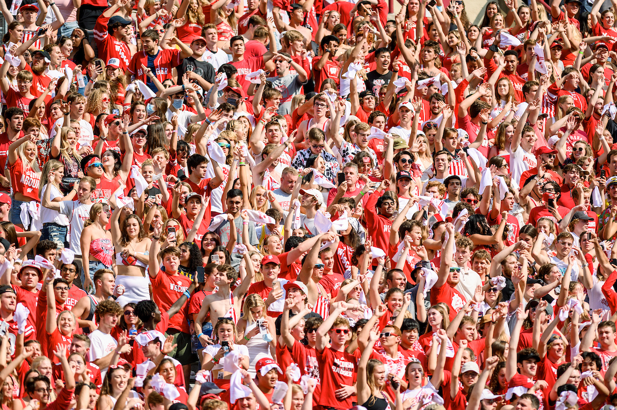 A crows of people wearing red and white cheer excitedly in the student section at a UW–Madison football game on a sunny day.