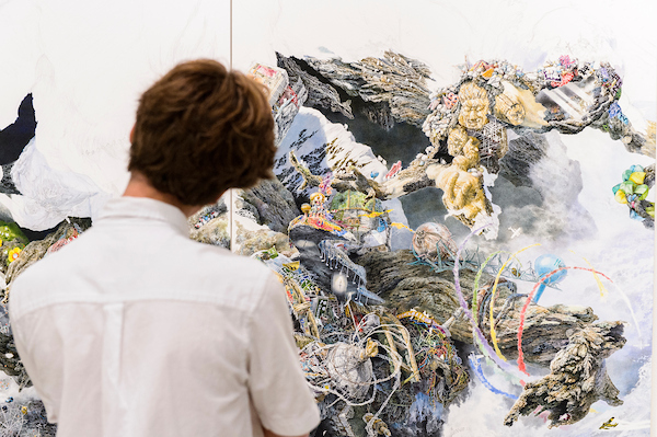 Person views a large mixed-media mural with abstract shapes and figures.