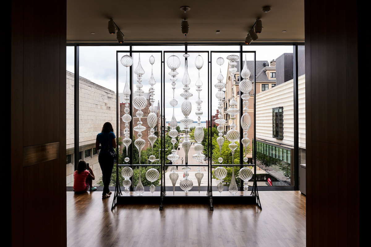 Hanging glass installation in front of a tall window, with two people looking on.