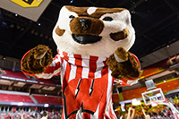 Bucky Badger excitedly pointing at the camera with fans in the background.